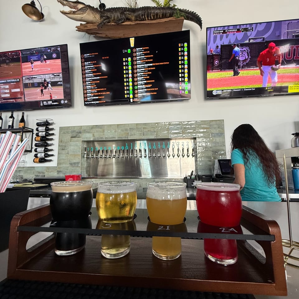 A four-glass beer flight in front of Chipley Station's taps and sports screens.